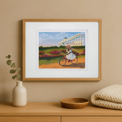 Framed artwork of a raccoon on a bicycle in a garden setting, displayed on a wooden shelf with decorative items.