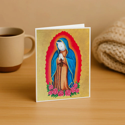 Card with a cat in a blue robe on a gold background, resembling a religious icon, placed on a wooden surface with a mug and fabric in the background.