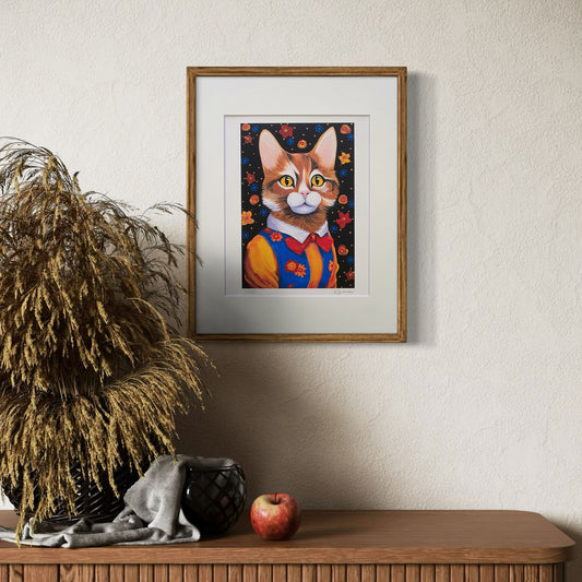 A framed print of Maisie the Cat hangs on a neutral-colored wall, adding a bold pop of color to the room. The print is matted with a white border and placed in a natural wood frame. Below it, a wooden console table holds a woven basket with dried plants, a folded gray cloth, a black ceramic vase, and a red apple, creating a cozy and artistic atmosphere.