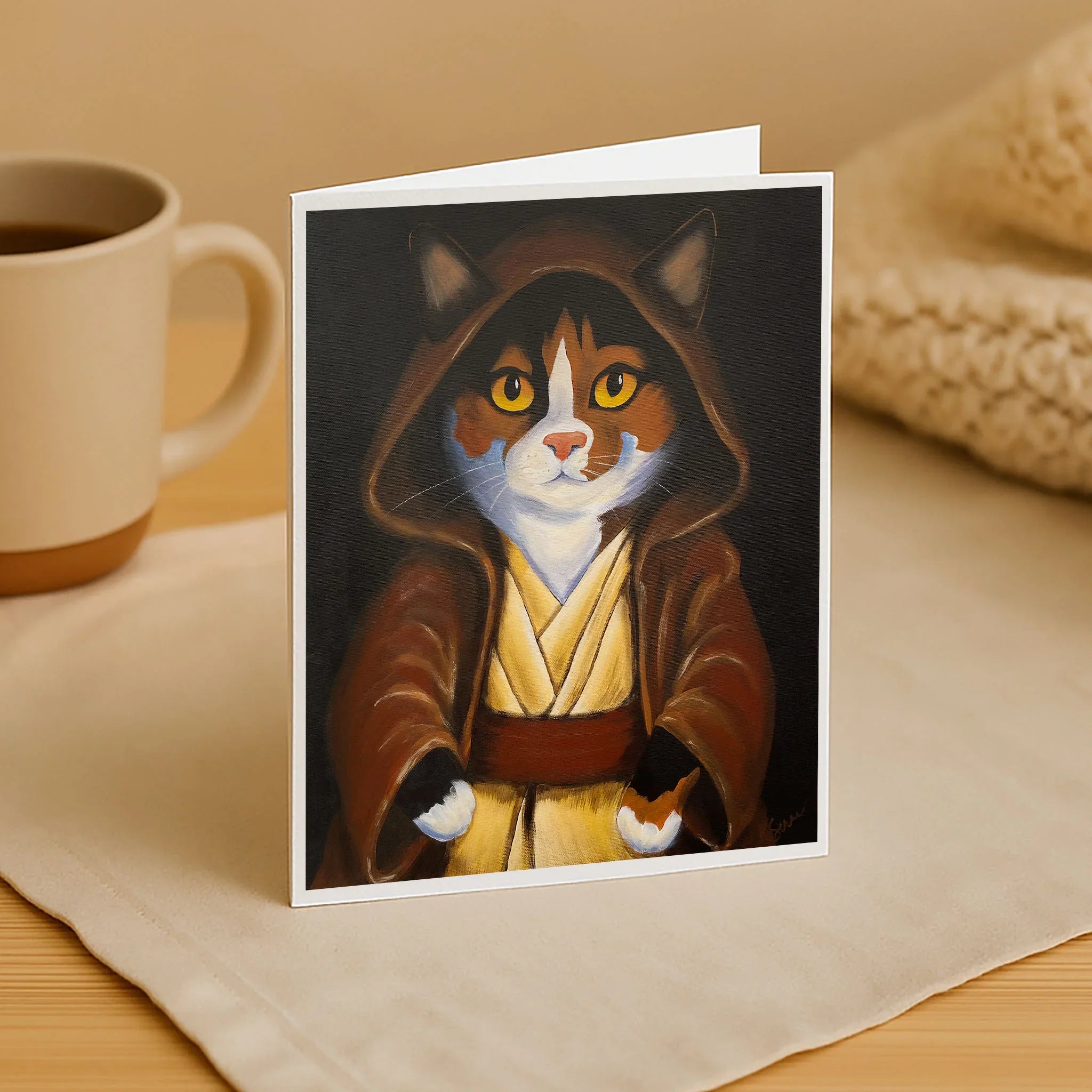 Greeting card with a cat in a robe on a table with a mug and blanket.