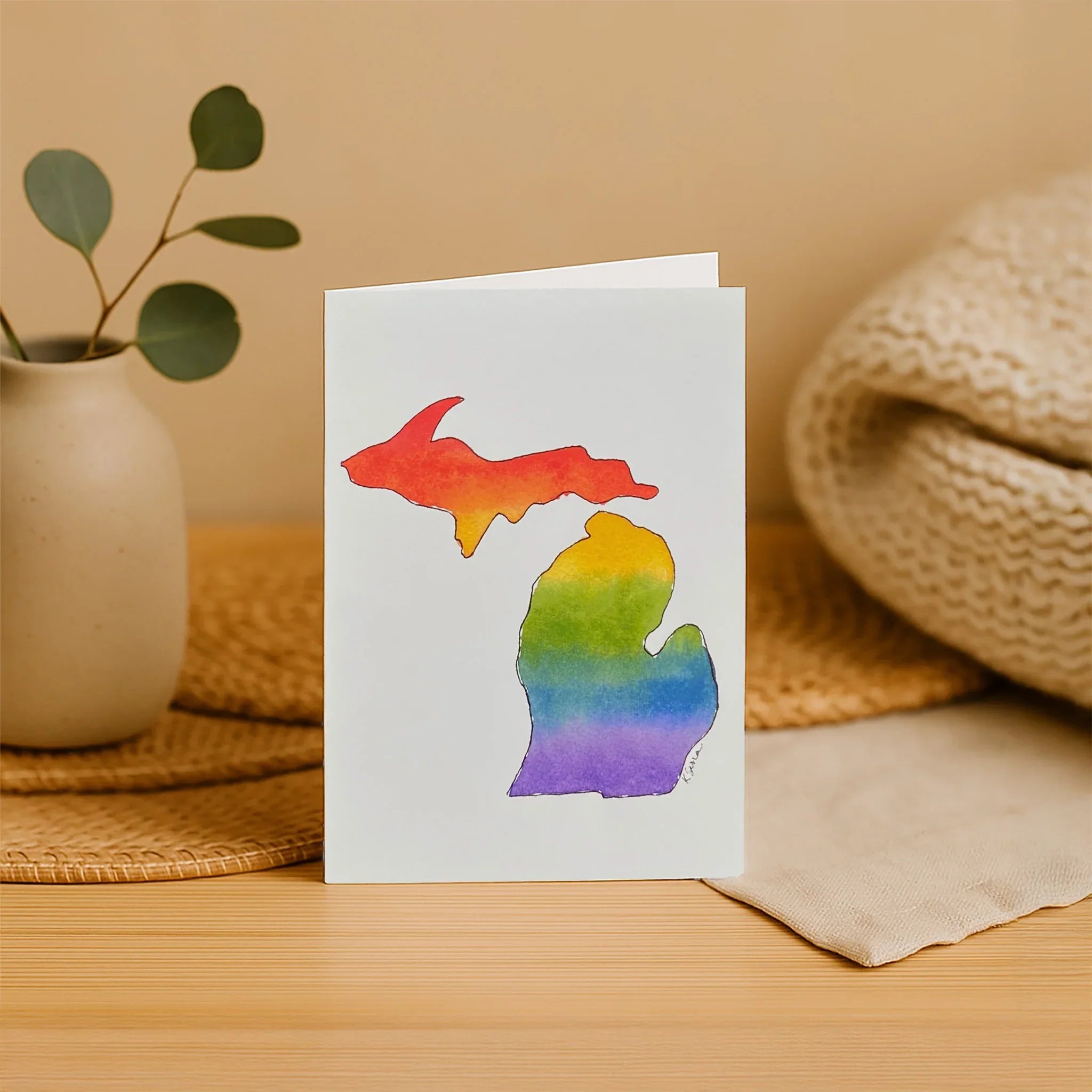 Card with a rainbow Michigan-shaped design on a wooden surface with a plant and woven mat in the background.