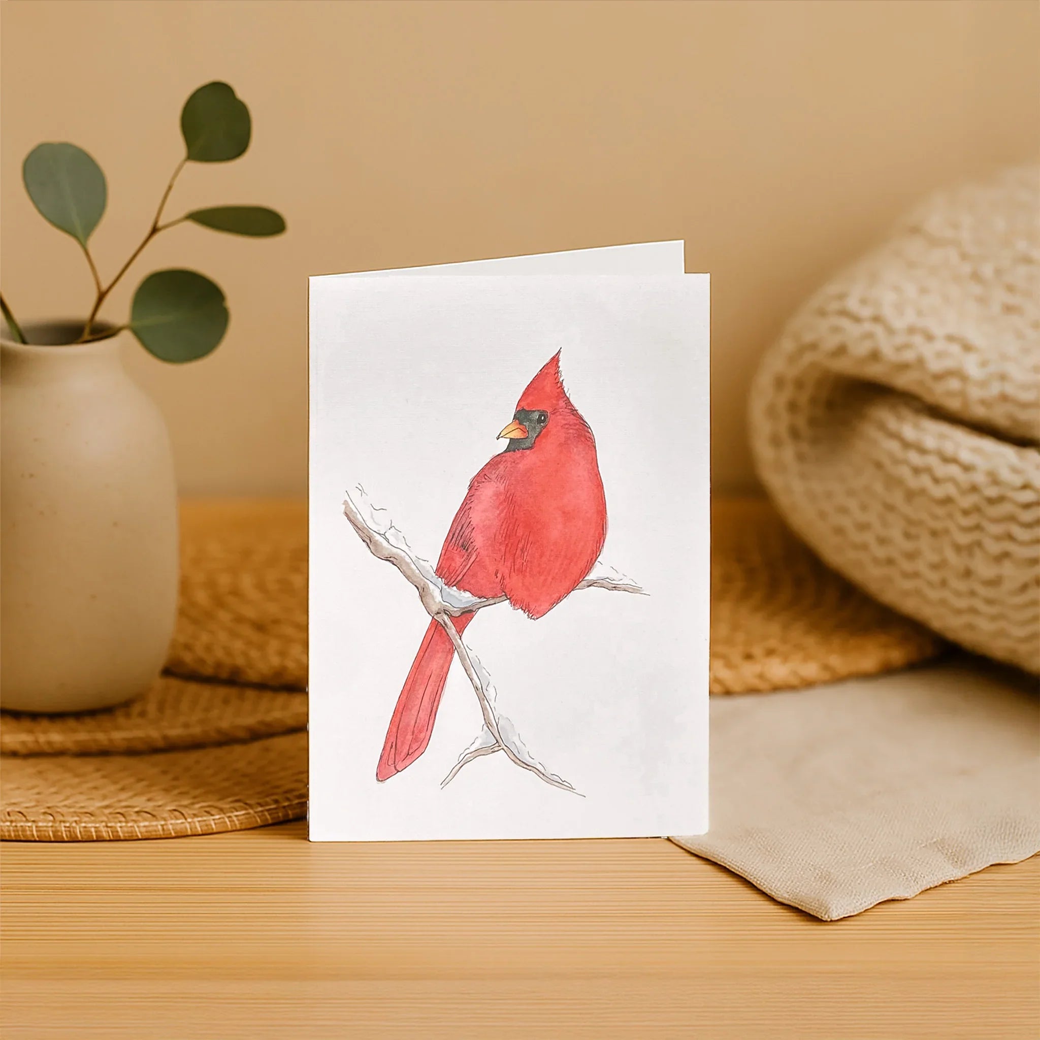 Card with a red bird illustration on a wooden surface with a plant and woven mat in the background.