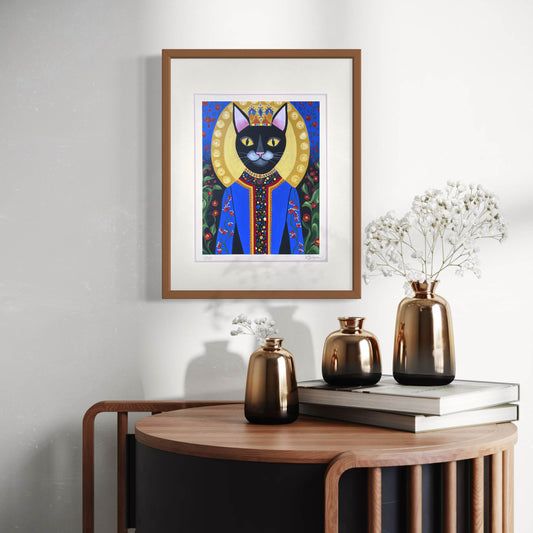 A framed print of Obi the Cat hangs on a neutral-colored wall, bringing a regal and majestic presence to the space. Obi, a sleek black cat with piercing yellow eyes, wears an ornate royal blue robe with intricate beadwork and embroidery, and a golden halo-like crown behind his head. Below, a modern wooden console is decorated with stacked books and a set of metallic bronze vases filled with delicate white flowers.