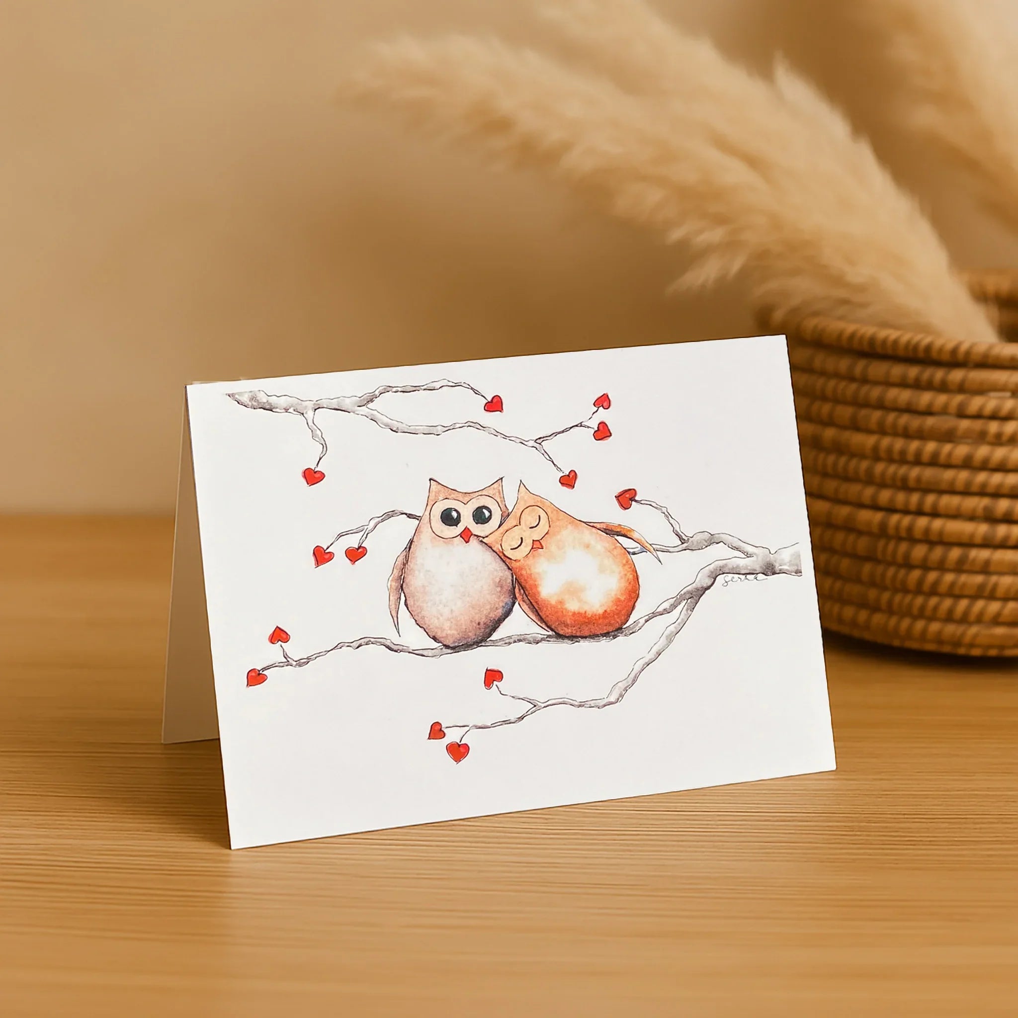 Greeting card with two owls on a branch and hearts, placed on a wooden surface with a woven basket in the background.