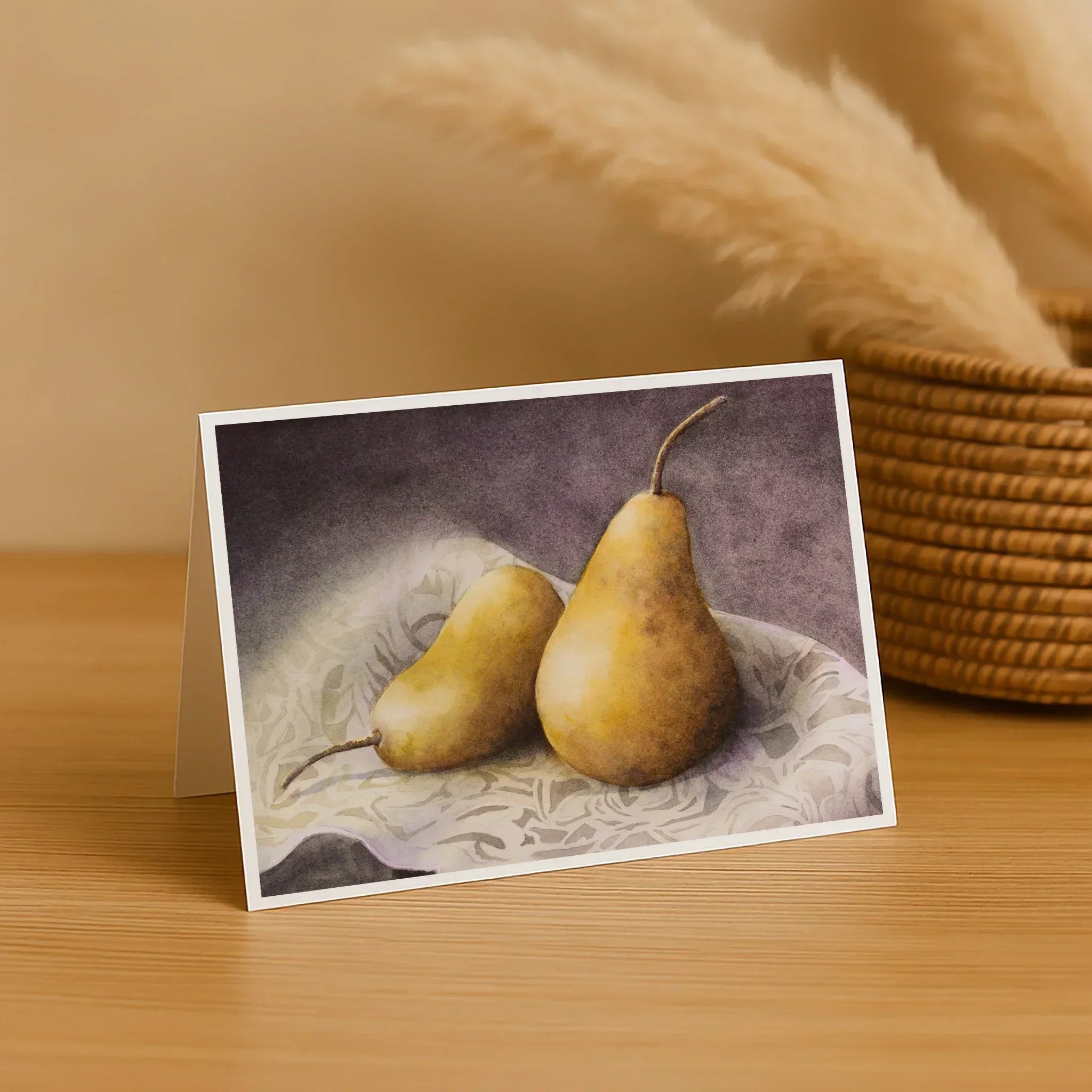 Greeting card with two pears on a textured surface, placed on a wooden table with a woven basket and decorative grasses in the background.