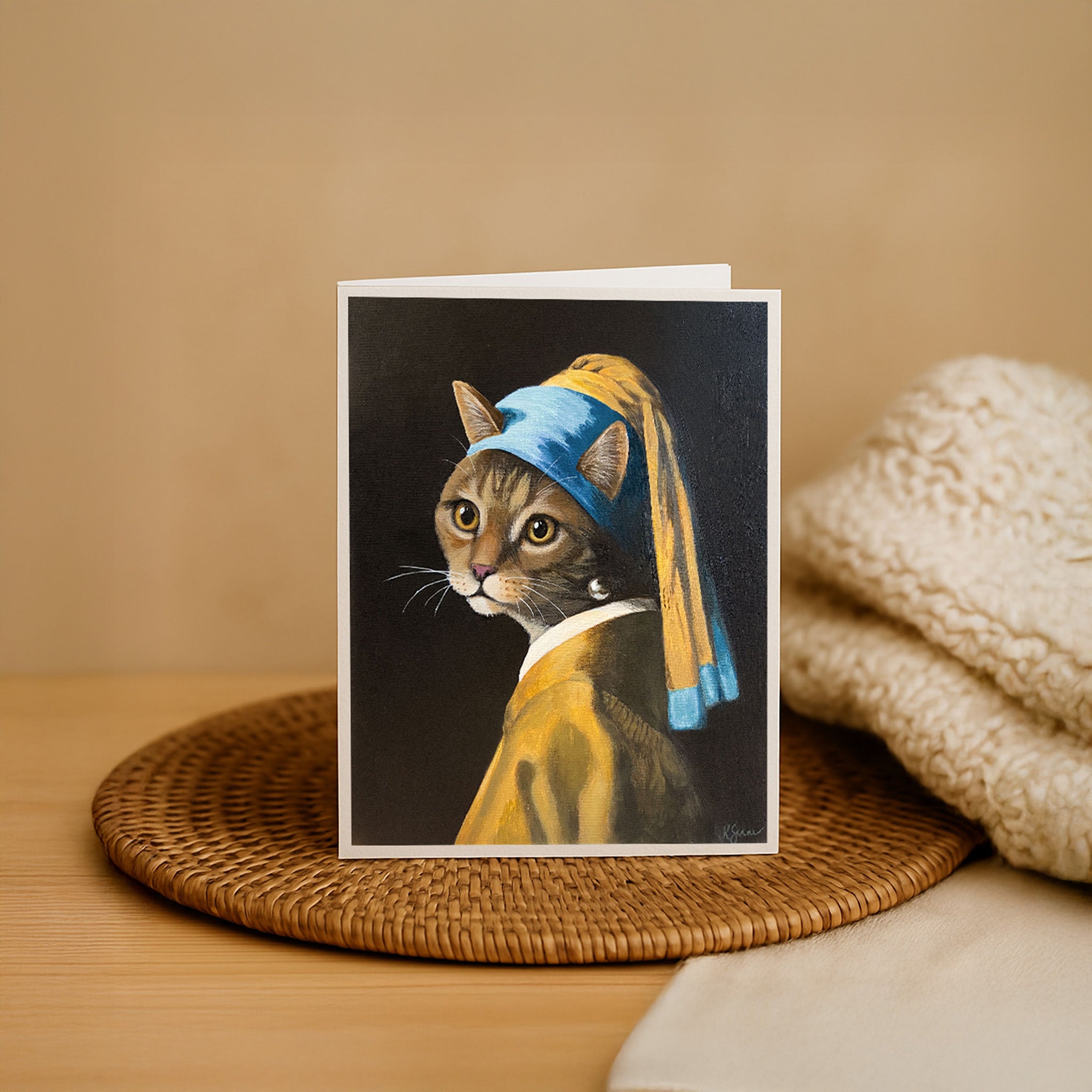 Card with a cat wearing a blue beret and yellow dress on a woven mat.