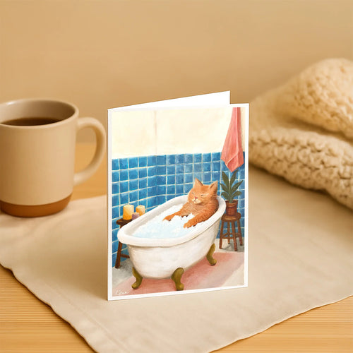 Greeting card with a cat in a bathtub on a table with a mug and blanket.