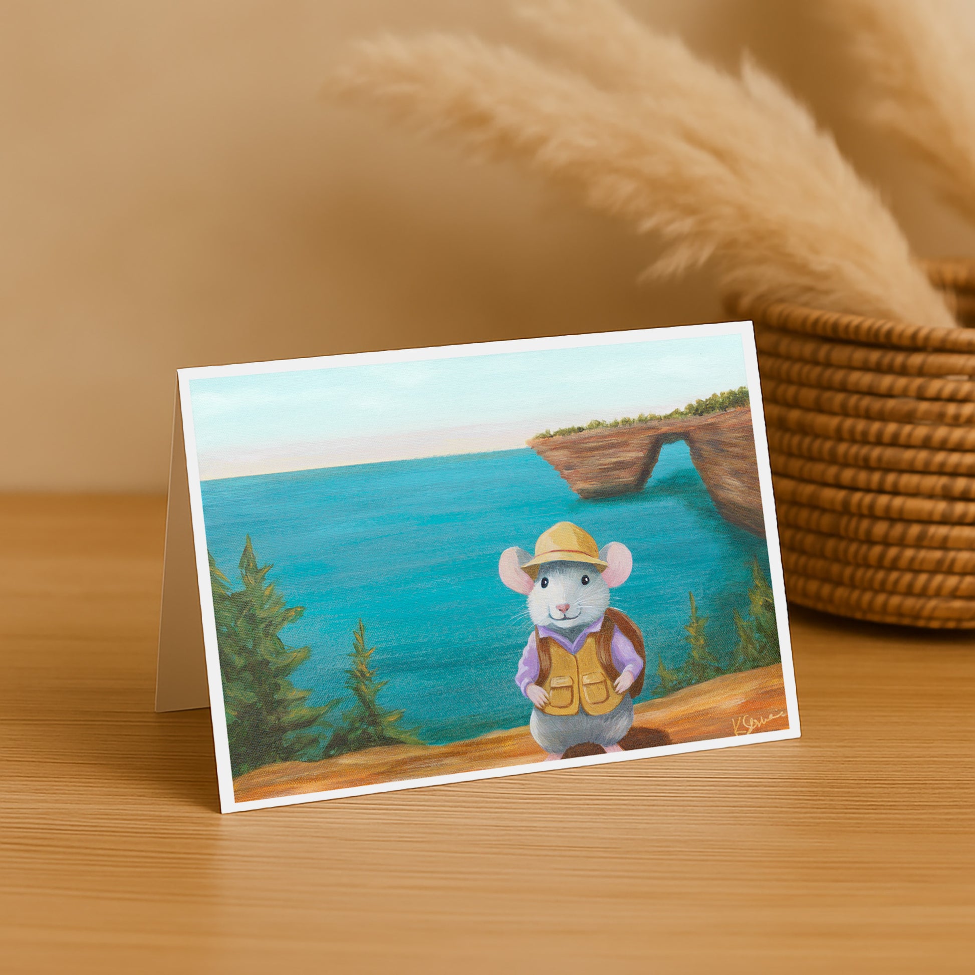 Greeting card with a mouse character by a lake on a wooden surface with a basket in the background.