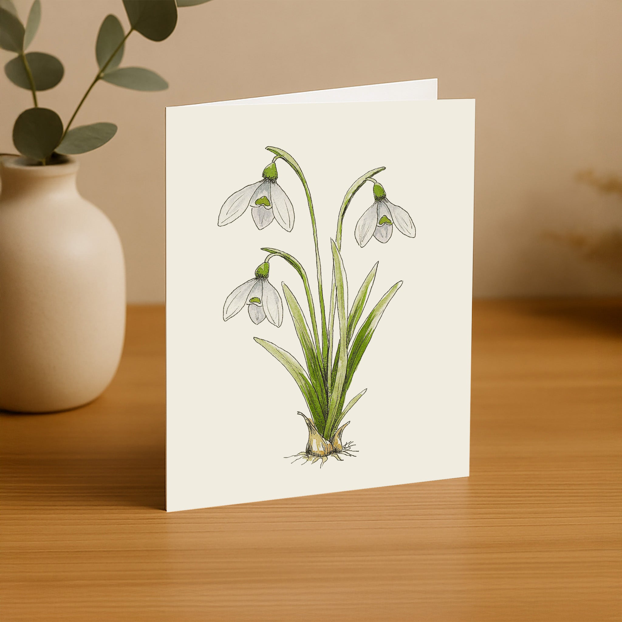 Greeting card with snowdrop illustration on a wooden surface with a vase in the background