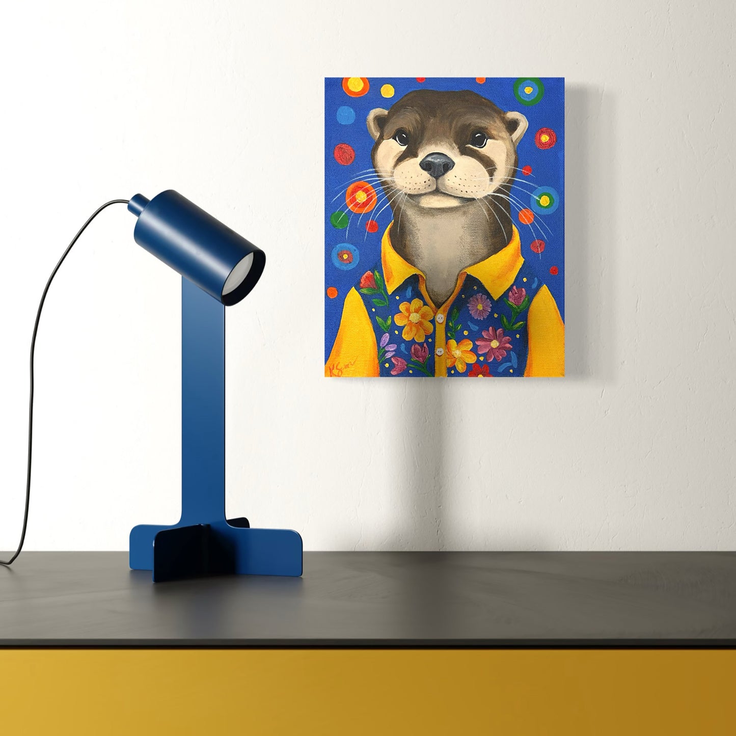 Canvas print of an otter wearing a yellow shirt with flowers on a white wall, next to a blue lamp.