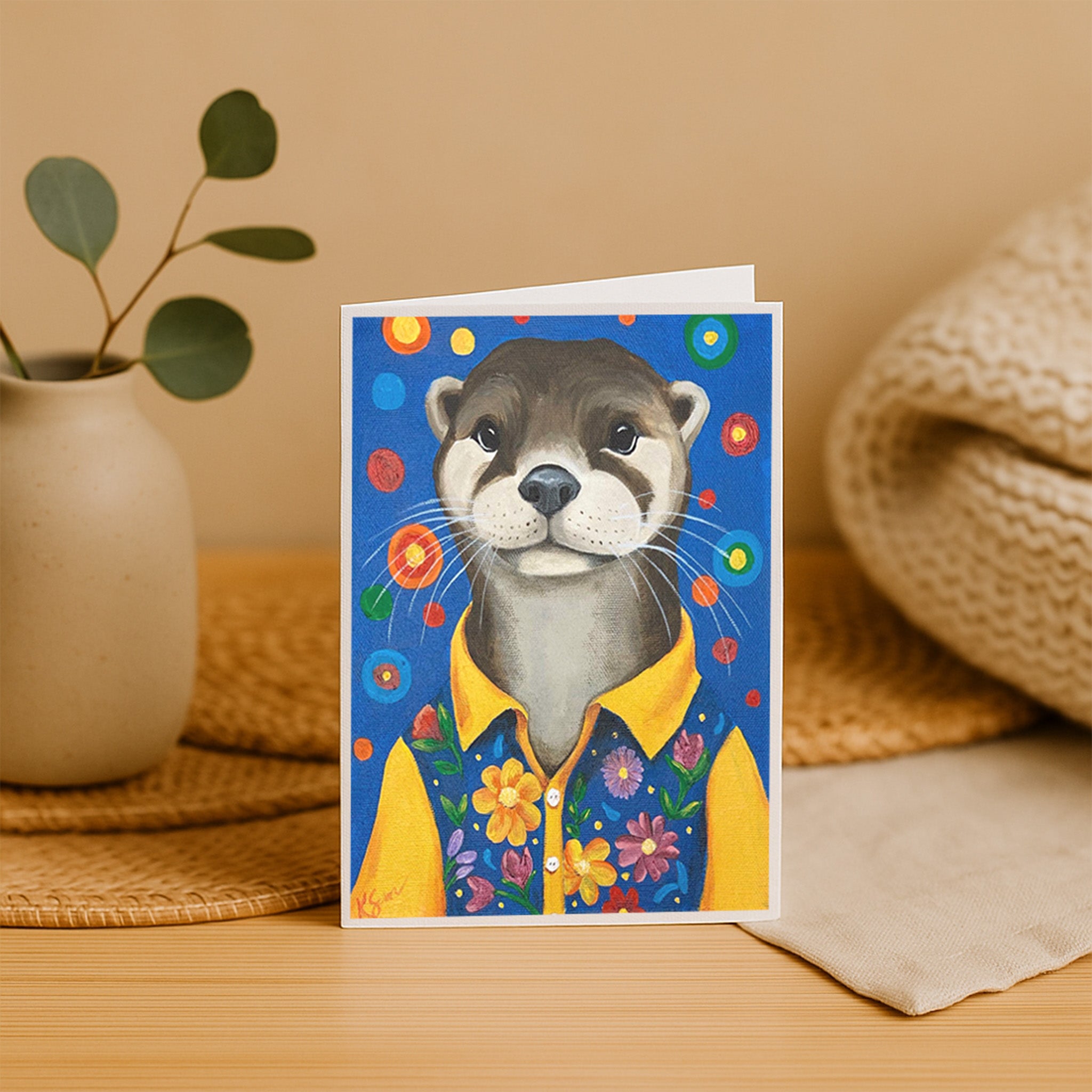 Greeting card with an otter illustration wearing a yellow shirt on a wooden surface.