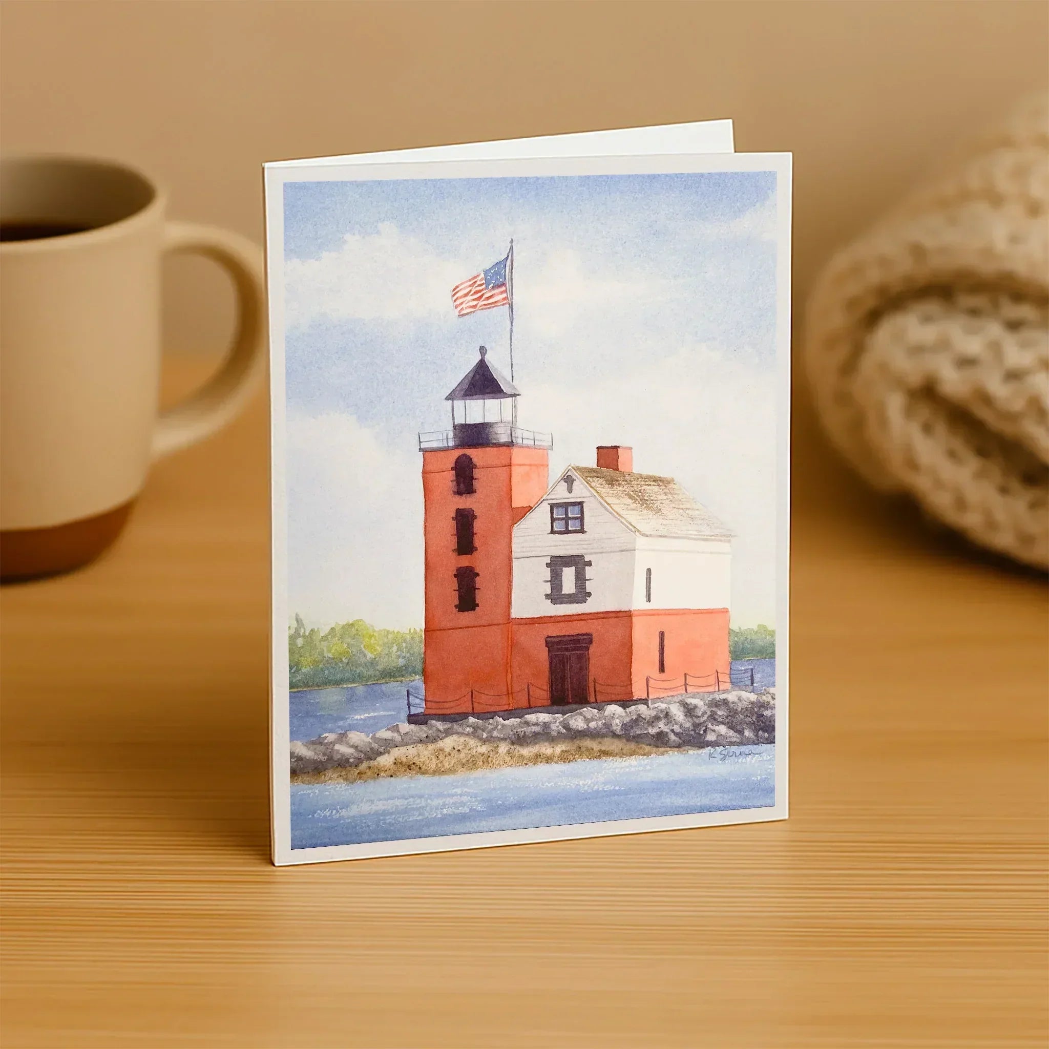 Greeting card with a lighthouse illustration on a wooden surface next to a mug.