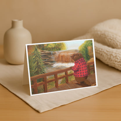 Greeting card with a bear in a checkered shirt looking at a waterfall, placed on a beige surface with a vase and fabric in the background.