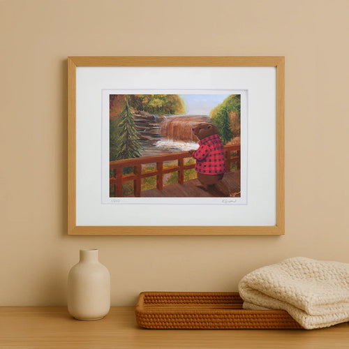 Framed artwork of a bear in a checkered shirt looking at a waterfall on a beige wall.