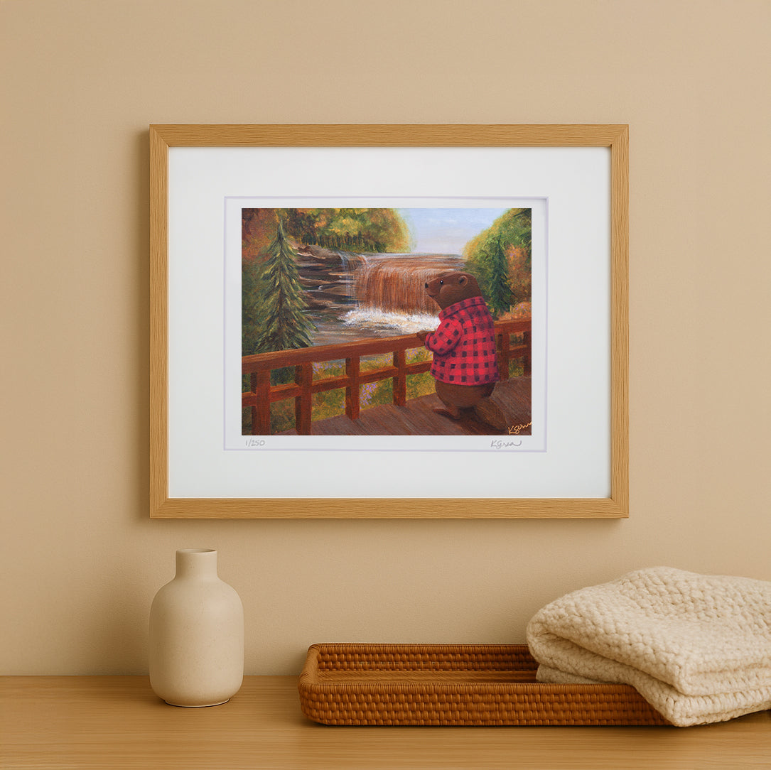 Framed artwork of a bear in a checkered shirt looking at a waterfall on a beige wall.