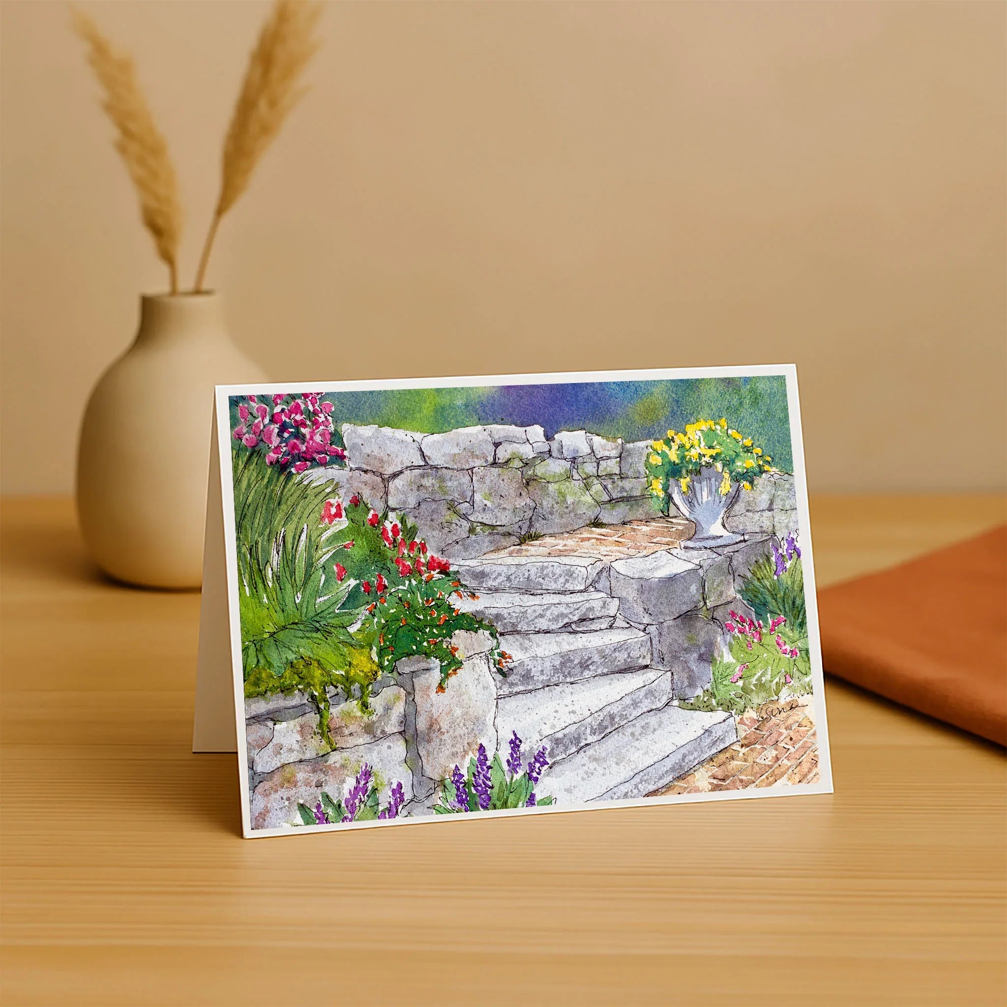 Greeting card with a garden scene featuring stone steps and flowers, placed on a wooden surface.