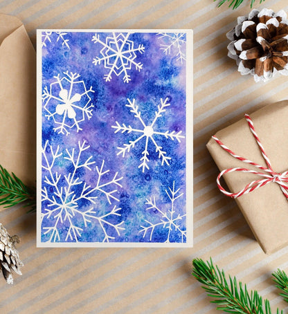 A watercolor painting of white snowflakes on a textured blue and purple background, creating a wintry abstract design.