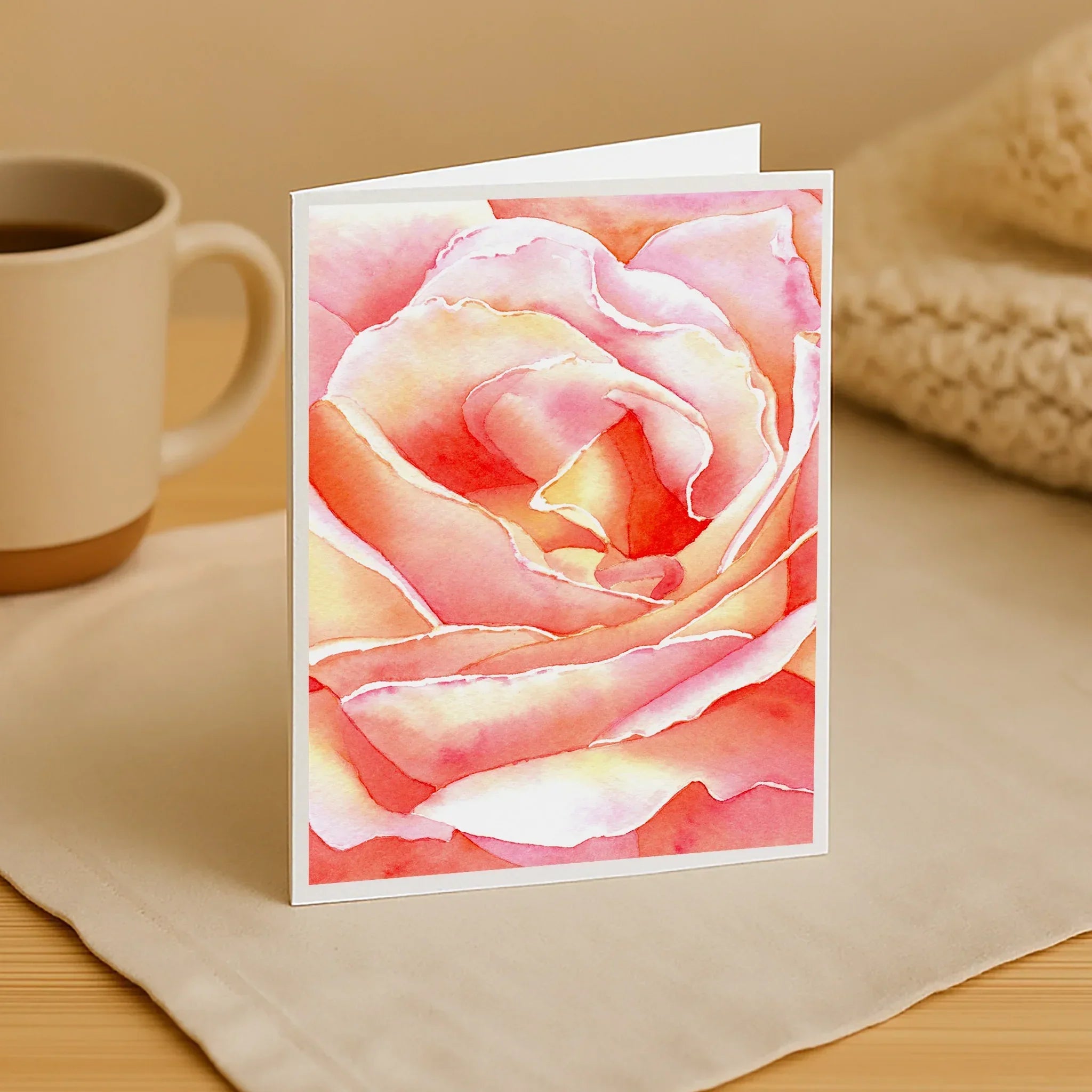 Greeting card with a pink rose design on a wooden surface with a mug and fabric.