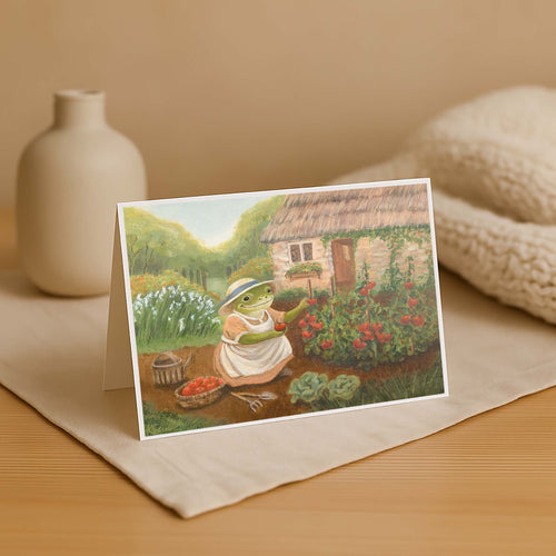 Greeting card with a frog character in a garden on a wooden surface with a vase and fabric in the background.