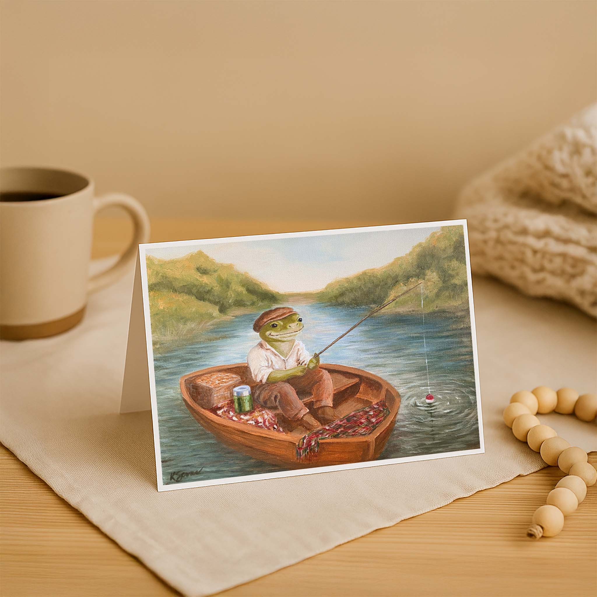 Greeting card with a frog in a boat fishing, placed on a wooden surface with a mug and beads.