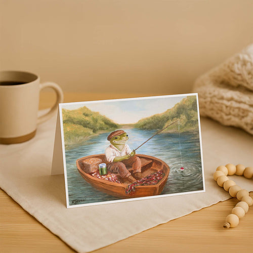 Greeting card with a frog in a boat fishing, placed on a wooden surface with a mug and beads.