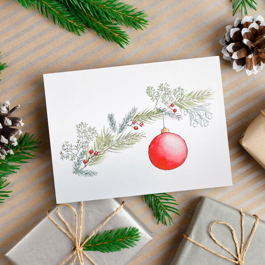 A watercolor and ink painting of a red Christmas ornament hanging from a branch of pine and holly with red berries.