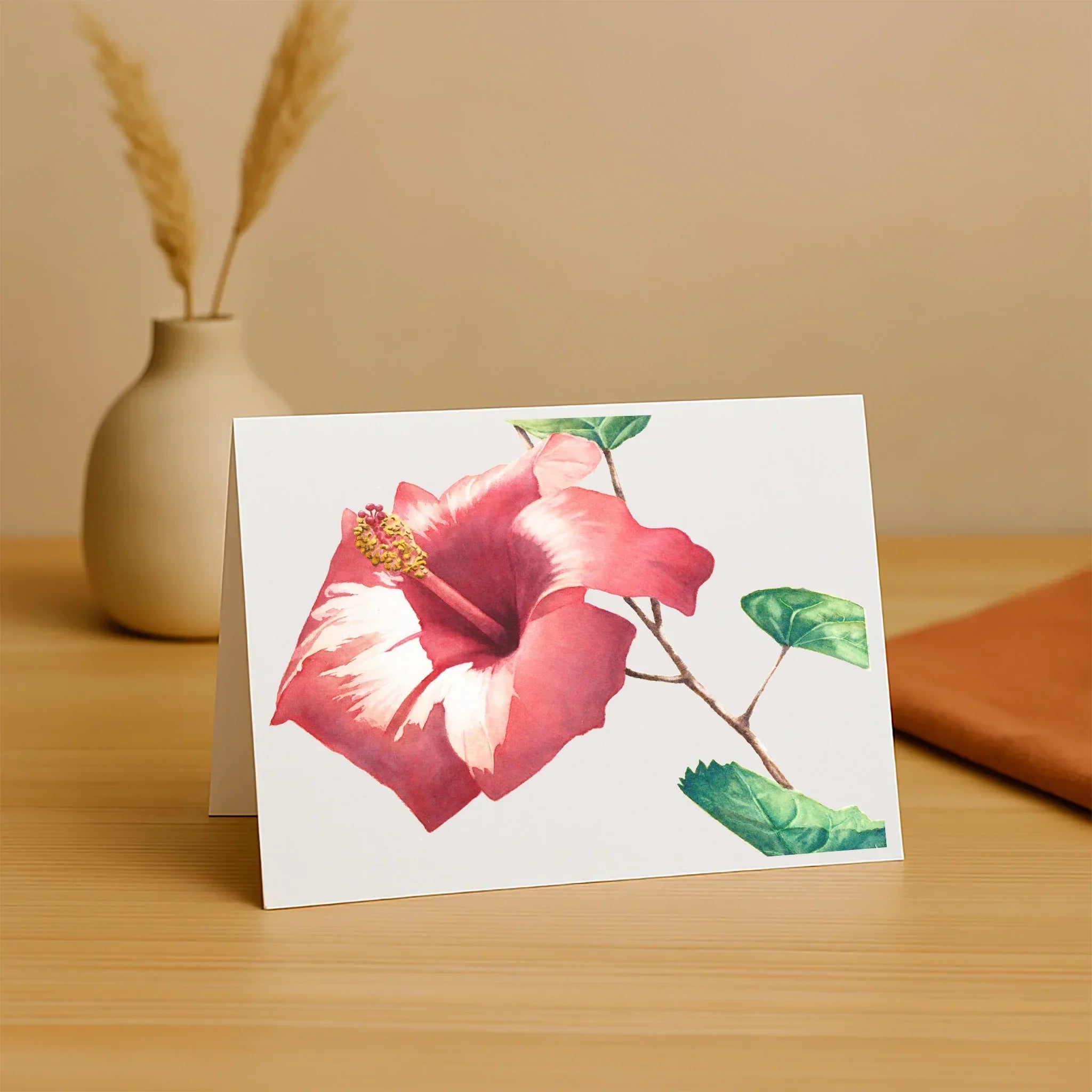 Greeting card with pink flower design on a wooden surface with a vase in the background