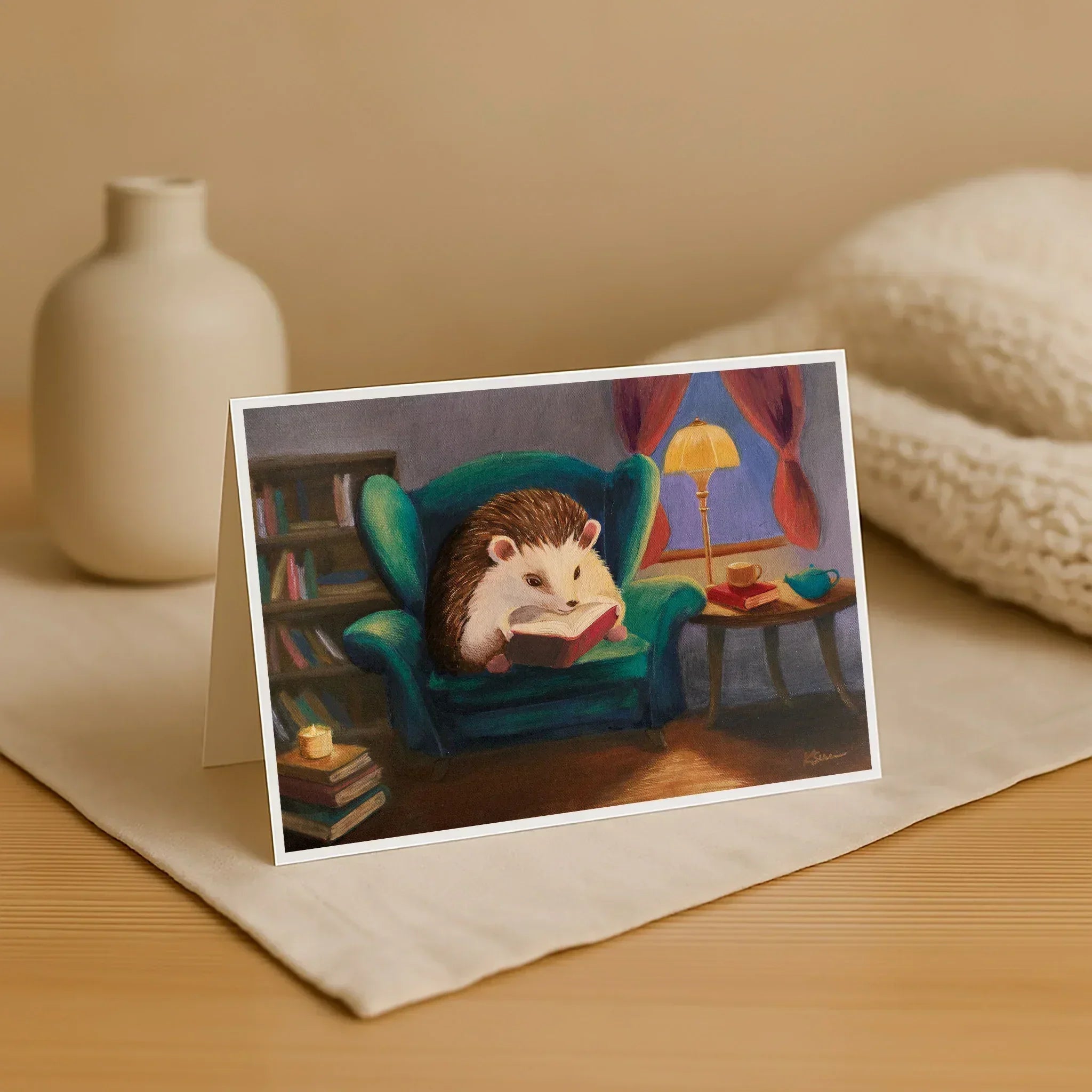 Greeting card with a hedgehog reading a book in an armchair, placed on a surface with a vase and blanket in the background.