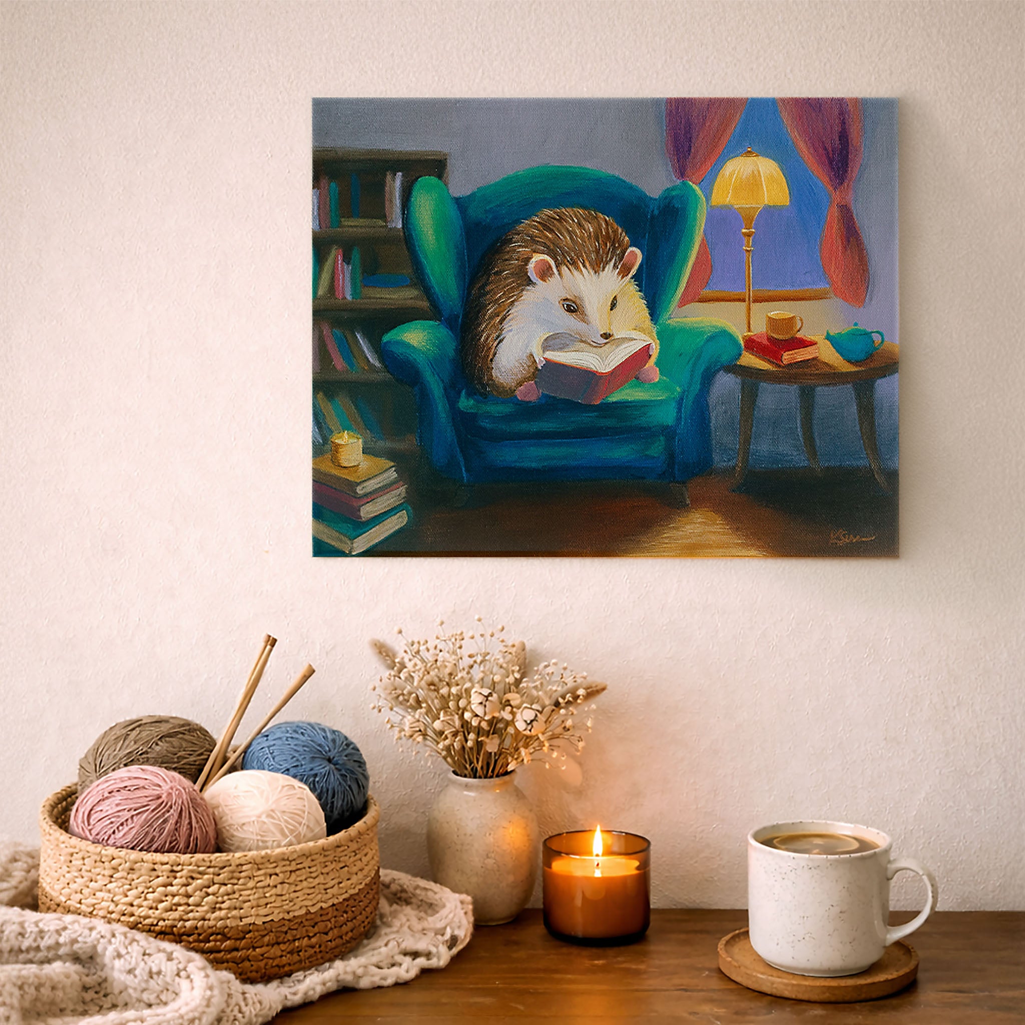 Painting of a hedgehog reading a book on an armchair, displayed above a cozy table setting with yarn, a mug, and a candle.