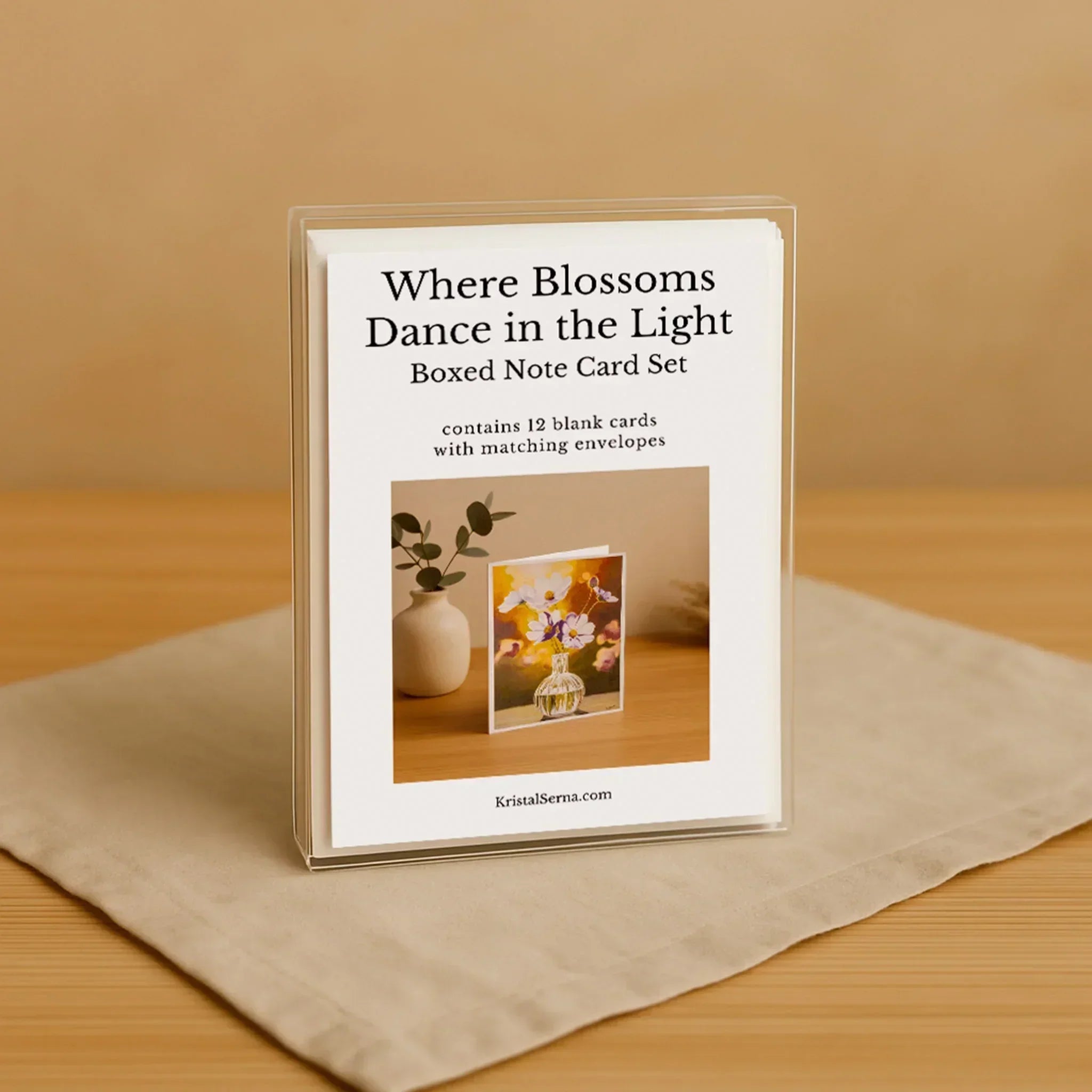 Boxed note card set titled 'Where Blossoms Dance in the Light' on a beige surface with a wooden background.