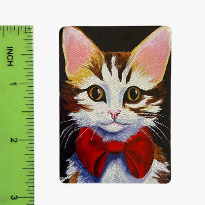 Painting of a cat with a red bow tie next to a ruler for scale.