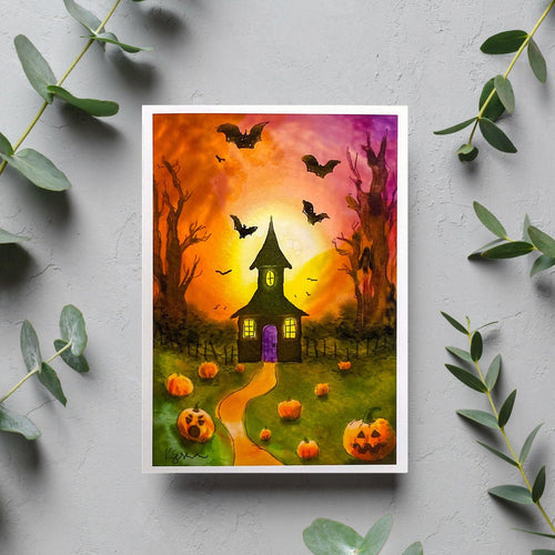 A greeting card with an artistic depiction of a house at dusk with bats flying, pumpkins on the ground, and a glowing orange sky in the background, presented on a bed of decorative green leaves.